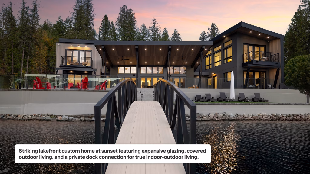 A modern lakefront custom home at sunset featuring expansive glazing, dark grey and wood siding, and glass-walled balconies. A central covered outdoor living area with recessed lights sits above a tiered patio furnished with red Adirondack chairs and lounge chairs. A private bridge with black railings connects the home to the water, all set against a backdrop of tall evergreen trees and a pink-hued sky.
