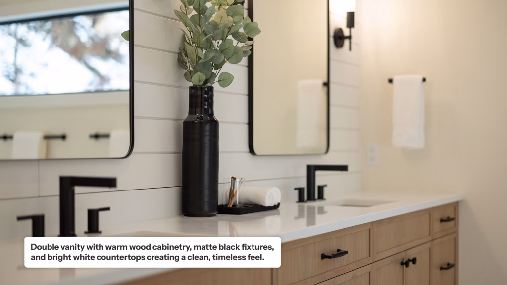 A modern bathroom double vanity featuring warm light-wood cabinetry, white countertops, and matte black faucets. Two rectangular, black-framed mirrors are mounted on a white shiplap wall, with a tall black vase of eucalyptus branches centered between the sinks.