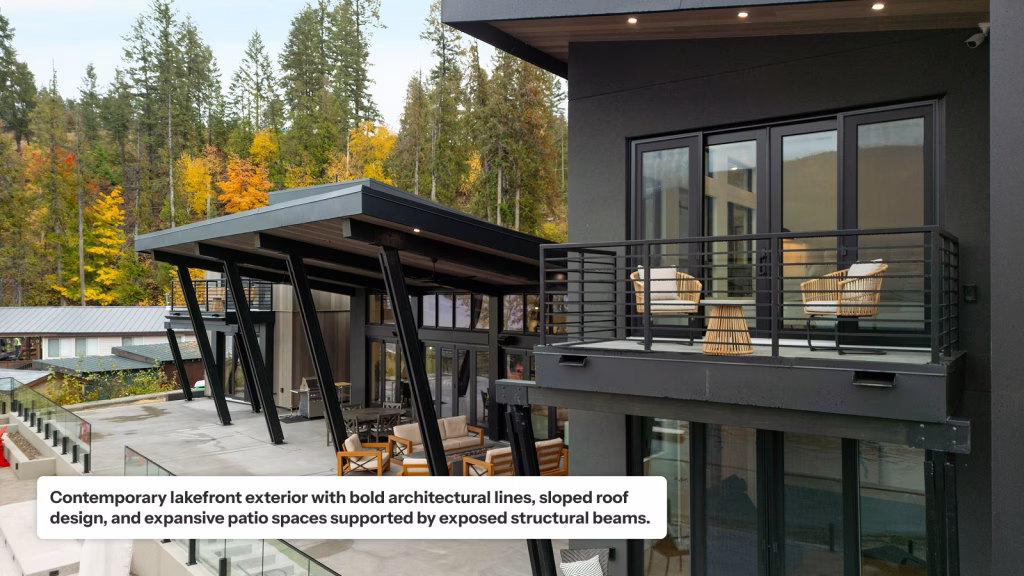 A contemporary lakefront home exterior featuring bold architectural lines and a sloped roof supported by angled black structural beams. The scene includes a large concrete patio with outdoor seating and a modern upper-level balcony with black metal railings, overlooking a forest with vibrant autumn foliage.