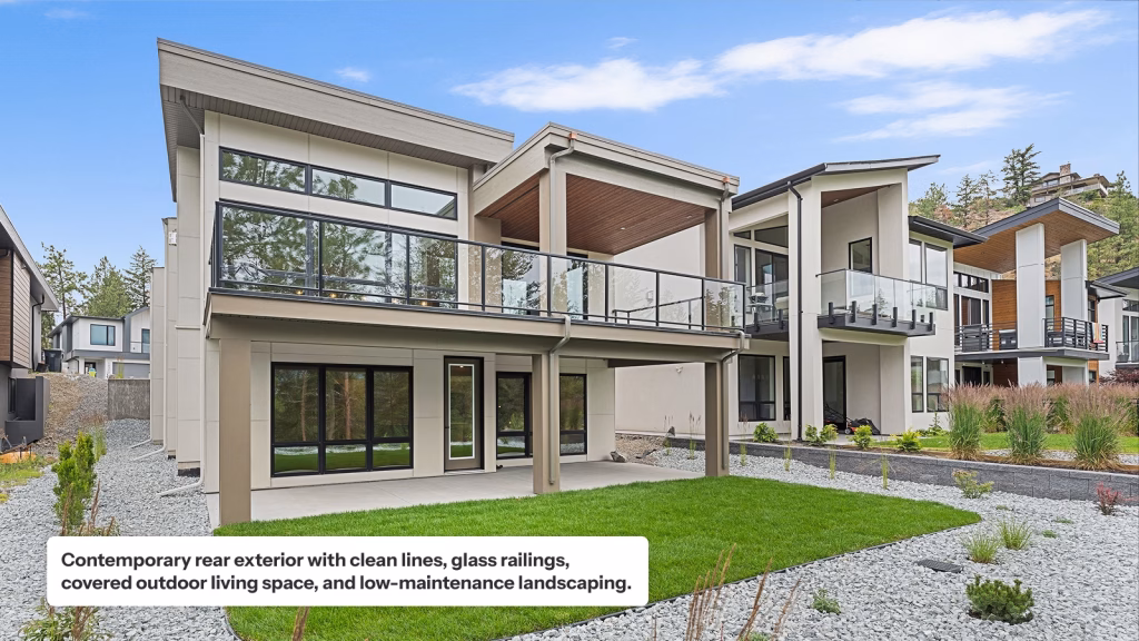 The rear exterior of a contemporary multi-story home featuring neutral-toned siding, clean lines, and large windows. It includes a spacious upper-level deck with glass railings and a wood-slat ceiling, overlooking a manicured green lawn surrounded by light grey gravel landscaping.