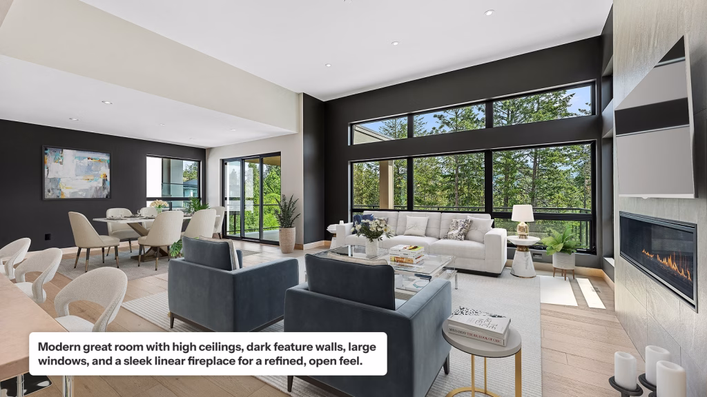 A spacious, modern great room featuring high ceilings, dark charcoal accent walls, and large windows with views of trees. The open layout includes a living area with a white sofa, blue armchairs, and a sleek linear fireplace, alongside a contemporary dining area in the background.