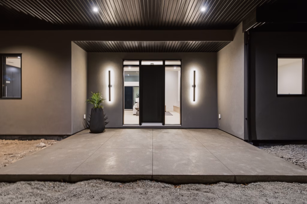 Covered entry of an Osoyoos custom home at night with linear sconces flanking a dark front door and glass sidelights.