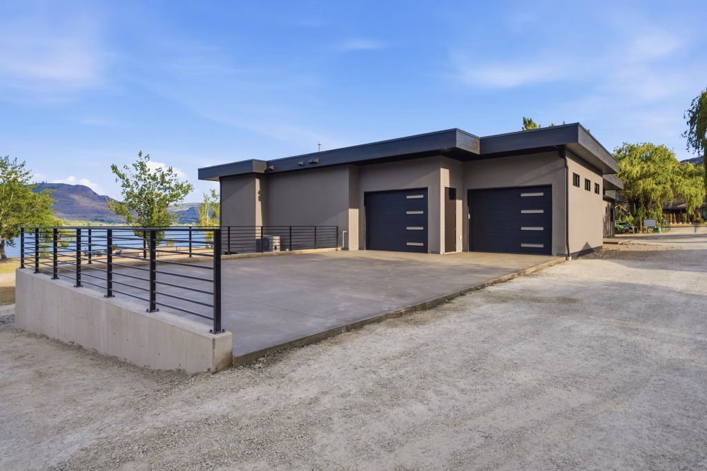 Two dark overhead garage doors and a concrete parking pad at this Osoyoos custom home, with the lake visible beyond.