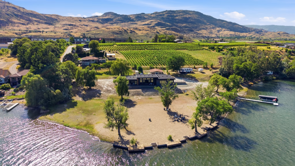 Daytime aerial of an Osoyoos lakefront home set behind a wide sandy beach, with vineyard rows and hills beyond.