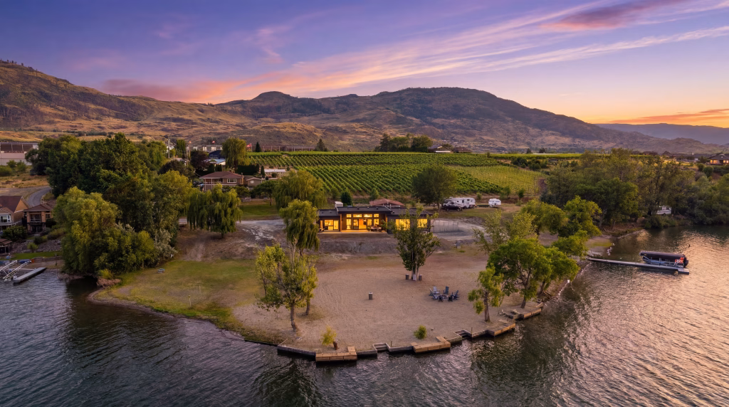 Twilight aerial of an Osoyoos lakefront home glowing above a sandy beach, with vineyard and mountains fading behind.