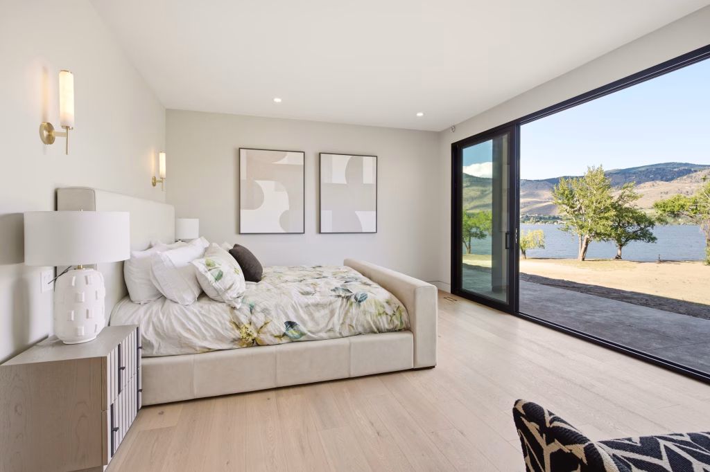Primary bedroom with gold-toned wall sconces and sliding glass doors framing the lake at this Osoyoos lakefront home.